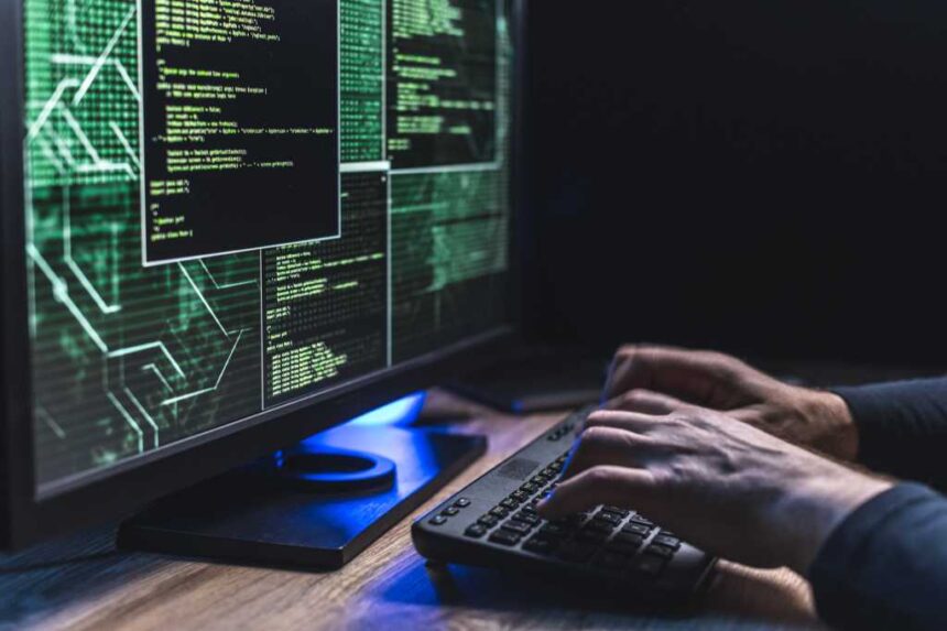 Hands use a keyboard at a desktop display showing lines of code in a dimly lit workspace.