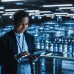 Handsome Smiling IT Specialist Using Tablet Computer in Data Center. Succesful Businessman and e-Business Entrepreneur Overlooking Server Farm Cloud Computing Facility.