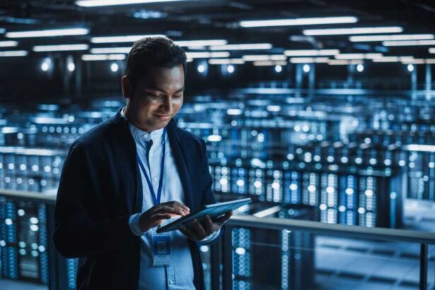 Handsome Smiling IT Specialist Using Tablet Computer in Data Center. Succesful Businessman and e-Business Entrepreneur Overlooking Server Farm Cloud Computing Facility.