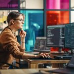 Female Junior Software Engineer Writes Code on Desktop Computer With Two Monitors and Laptop Aside In Stylish Office. Caucasian Woman Working On Artificial Intelligence Service For Big Tech Company.