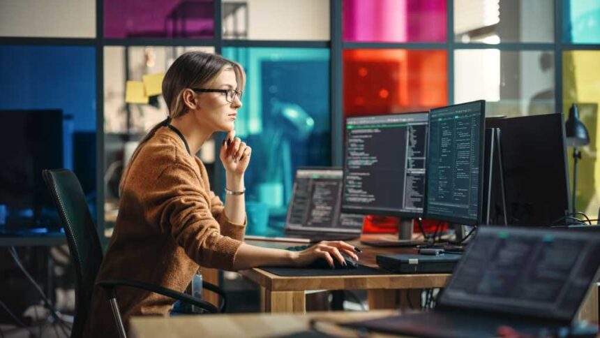 Female Junior Software Engineer Writes Code on Desktop Computer With Two Monitors and Laptop Aside In Stylish Office. Caucasian Woman Working On Artificial Intelligence Service For Big Tech Company.