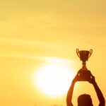 Winner holding golden trophy cup above head sun and golden sky in background