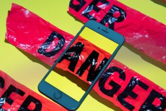 A smartphone sits on top of a surface with red tape reading “DANGER.” Where one strip intersects the phone, it continues inside the phone’s screen.