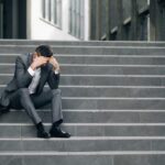 Fired unhappy european bearded man losing job sitting on stairs. Upset young businessman in frustration outside office department. Unemployment. Crisis.