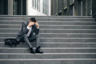 Fired unhappy european bearded man losing job sitting on stairs. Upset young businessman in frustration outside office department. Unemployment. Crisis.