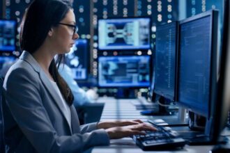 Female Engineer Controller Observes Working of the System. In the Background People Working and Monitors Show Various Information.