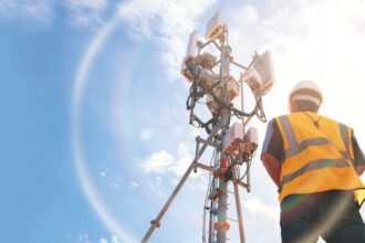 Helmeted asian male engineer works in the field with a telecommunication tower that controls cellular electrical installations to inspect and maintain 5G networks installed on high-rise buildings.