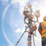 Helmeted asian male engineer works in the field with a telecommunication tower that controls cellular electrical installations to inspect and maintain 5G networks installed on high-rise buildings.