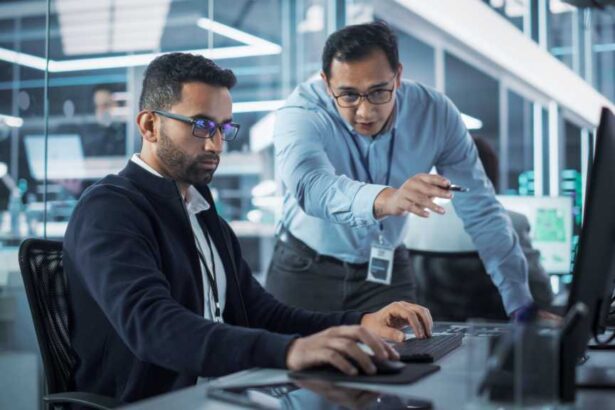 Male Project Supervisor Talks to an Industry 4 Engineer Who Works on Desktop Computer in Robotics Startup Industrial Office. Software Developer Discussing Work Related Tasks During a Meeting.