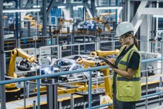 Female Car Factory Engineer in High Visibility Vest Using Tablet Computer. Automotive Industrial Manufacturing Facility Working on Vehicle Production with Robotic Technology. Automated Assembly Plant.