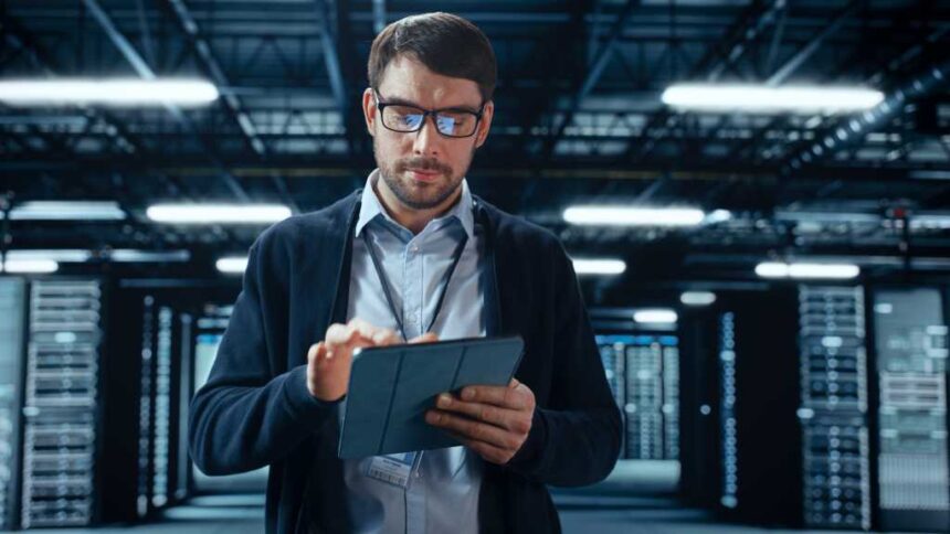 Male IT Specialist Walks Between Row of Operational Server Racks in Data Center. Engineer Uses Tablet Computer for Maintenance. Concept for Cloud Computing, Artificial Intelligence, Cybersecurity.