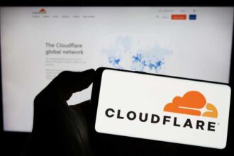 A photograph of a person holding a smartphone with the Cloudflare logo displayed. Behind the phone is a blurred monitor displaying the Cloudflare website.,