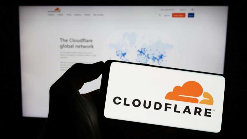 A photograph of a person holding a smartphone with the Cloudflare logo displayed. Behind the phone is a blurred monitor displaying the Cloudflare website.,
