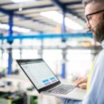 Side view of technician or engineer with headset and laptop standing in industrial factory.