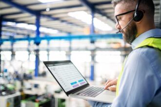 Side view of technician or engineer with headset and laptop standing in industrial factory.