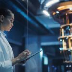 Female Engineer Working on Quantum Computer Hardware, Using Tablet Computer to Access Technical Specifications and Control Processor Parameters. Group of Developers Work on AI Behind a Desk